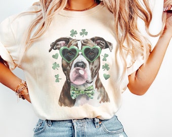 Op de afbeelding: Een wit T-shirt met een bruine en witte hond die een groene hartvormige zonnebril en een groene strik draagt. De hond is omringd door groene klavertjes.
