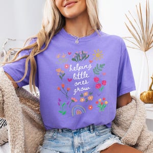 May include: A purple t-shirt with a floral design and the text "helping little ones grow".