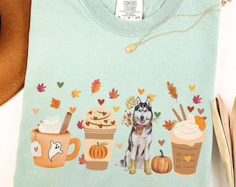 May include: A light green Comfort Colors t-shirt featuring a fall-themed design. The design includes a cartoon husky, pumpkins, and coffee cups with whipped cream. The shirt also has autumn leaves and the word "BAY" printed on the top.