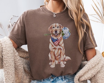 Puede incluir: Una camiseta marrón con un gráfico de un perro golden retriever que lleva una bandana con el diseño de la bandera estadounidense y sostiene un ramo de flores azules y blancas en su boca.