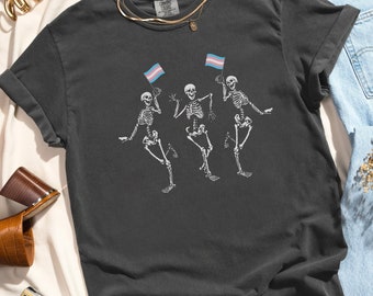 May include: A dark gray t-shirt with a graphic of three skeletons dancing and holding transgender pride flags. The shirt has a distressed look and is made of a soft, comfortable material.