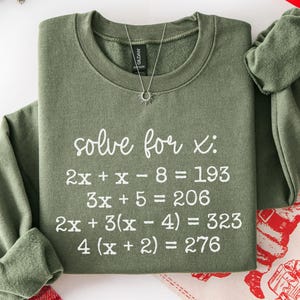 May include: A military green sweatshirt with the text "solve for x:" and several algebraic equations in white. The sweatshirt has long sleeves and a crew neck. A silver necklace with a sun pendant is visible.