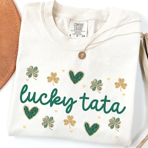 May include: A cream colored t-shirt with the text "lucky tata" in green lettering. The shirt is decorated with green shamrocks, gold glitter hearts, and gold glitter shamrocks. The shirt is labeled "Comfort Colors" and is available in ivory.