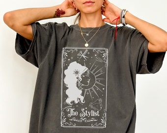 May include: An oversized t-shirt in a dark grey colour with a white graphic of a woman with a sun and moon design. The graphic also includes a pair of scissors, a hairdryer, and the text "The Stylist".