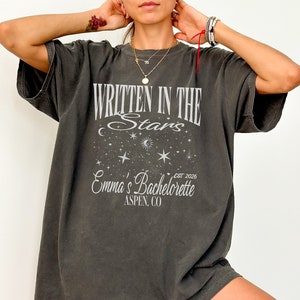 May include: A dark gray oversized t-shirt with a white graphic design that says "Written in the Stars" with a crescent moon, stars, and the text "Emma's Bachelorette Est. 2026 Aspen, CO".