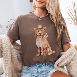 Peut inclure: T-shirt couleur expresso avec une illustration à l'aquarelle d'un golden doodle portant un chapeau et des bottes de cow-boy, tenant des marguerites. Le t-shirt a une coupe décontractée et est associé à un short en jean.