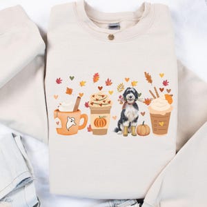May include: Sand-colored sweatshirt featuring a fall-themed design. The graphic includes a dog, pumpkins, and coffee cups with whipped cream. Colorful leaves and hearts are scattered around the design. The sweatshirt is folded on a light-colored surface.
