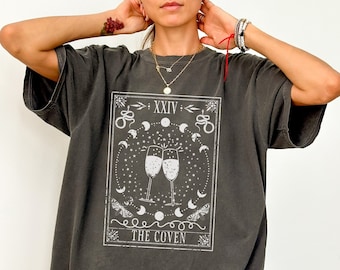 Pode incluir: T-shirt cinzenta escura com um design de carta de tarot com duas taças de champanhe, luas, estrelas e o texto "THE COVEN". A t-shirt tem um corte grande.