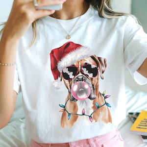 May include: White t-shirt featuring a Boxer dog wearing a Santa hat and sunglasses, blowing a bubblegum bubble. The dog is adorned with a string of colorful Christmas lights. A fun, festive design.
