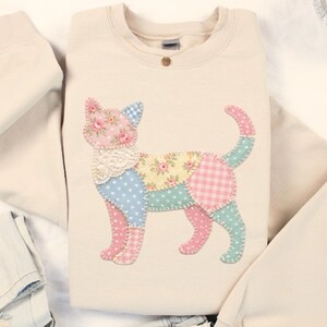 May include: A sand-coloured sweatshirt with a patchwork cat design. The cat is made from various patterned fabrics, including floral, polka dot, and gingham in pastel colours. The sweatshirt is folded with a pair of light-wash jeans.