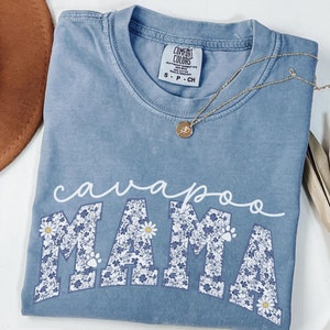 May include: A light blue t-shirt with a floral print design that spells out "cavapoo mama".