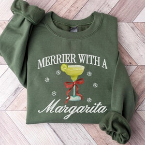 May include: Green Christmas sweatshirt with the text "Merrier with a Margarita" and a margarita glass with a lime wedge and a red bow.