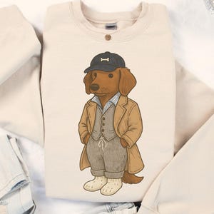 May include: Sand-colored sweatshirt featuring a cartoon dachshund wearing a cap with a bone, a coat, and a vest. The dog is depicted in a stylish outfit, with a light blue striped shirt and gray pants. The word "SAND" is printed at the bottom.
