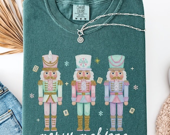 May include: A teal-colored sweatshirt with a graphic of three nutcrackers in pastel colors. The words "merry mahjong" are printed below the nutcrackers. A silver necklace is draped across the shirt.