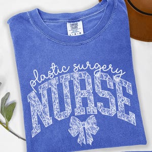 May include: A blue t-shirt with the text "Plastic Surgery Nurse" in white lace lettering. The text is in a large, bold font and is centered on the shirt. There is a white lace bow below the text. The shirt is a Comfort Colors brand and is size L-G.