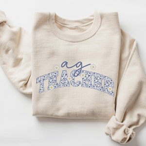 May include: A beige sweatshirt with the text "a g TEACHER" in blue with a daisy pattern.