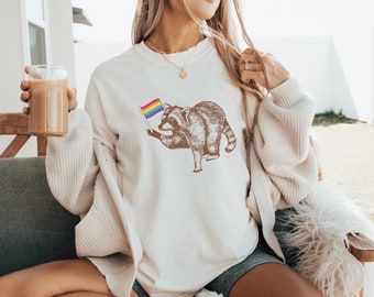 May include: A white sweatshirt with a graphic of a raccoon holding a rainbow flag. The raccoon is brown and has a black nose and eyes. The sweatshirt is worn by a person sitting on a white fluffy chair.