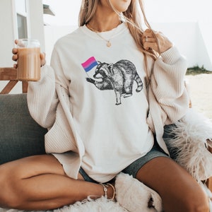 May include: A white crew neck sweatshirt with a graphic of a raccoon holding a bisexual pride flag. The raccoon is black and white and the flag is blue, pink, and purple.