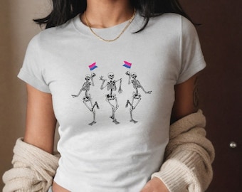 May include: A white cropped t-shirt with a graphic of three skeletons dancing. Each skeleton is holding a small flag with the bisexual pride colors.