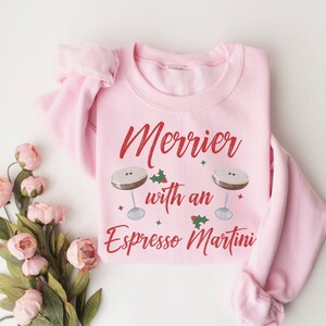 May include: Pink sweatshirt with the text "Merrier with an Espresso Martini" in red lettering. Two martini glasses with coffee and holly are on either side of the text.