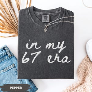 May include: A dark gray t-shirt with the text "in my 67 era" in white script. A silver necklace with a pendant rests on the shirt. The shirt is folded next to a pair of blue jeans and a beige sweater.