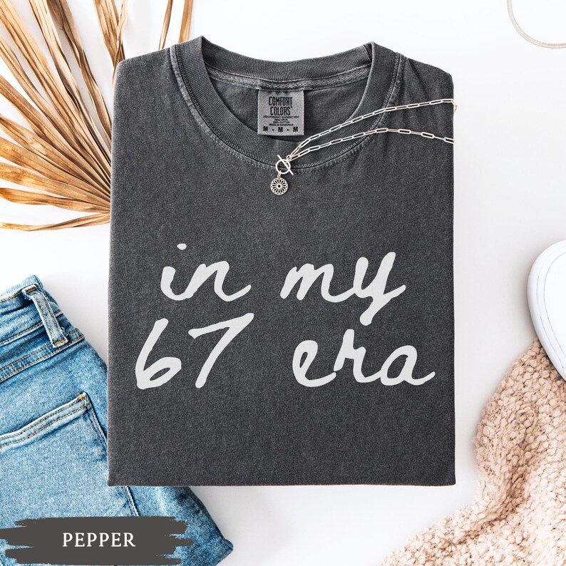 May include: A dark gray t-shirt with the text "in my 67 era" in white script. A silver necklace with a pendant rests on the shirt. The shirt is folded next to a pair of blue jeans and a beige sweater.