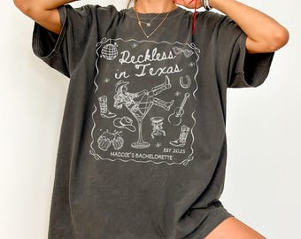 May include: A dark gray oversized t-shirt with a white graphic design that says "Reckless in Texas" with a cowboy boot, horseshoe, and martini glass. The graphic also includes the text "Maddie's Bachelorette est 2025".