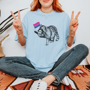 May include: A light blue t-shirt with a graphic of a raccoon holding a bisexual pride flag. The person wearing the shirt is sitting with their legs crossed and is making a peace sign with both hands.