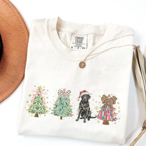 May include: A cream colored t-shirt with a Christmas themed design featuring three decorated Christmas trees and a black Labrador Retriever wearing a Santa hat. The trees are decorated with bows, ornaments, and lights. The text "COMFORT COLORS" is visible on the shirt.