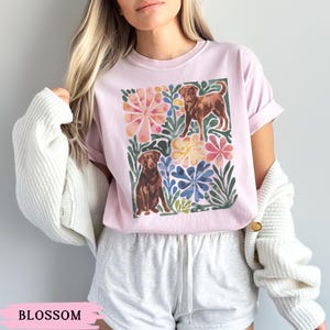 May include: A pale pink t-shirt with a watercolour design of two brown dogs amidst colourful flowers and green leaves. The word "BLOSSOM" is printed at the bottom of the image.