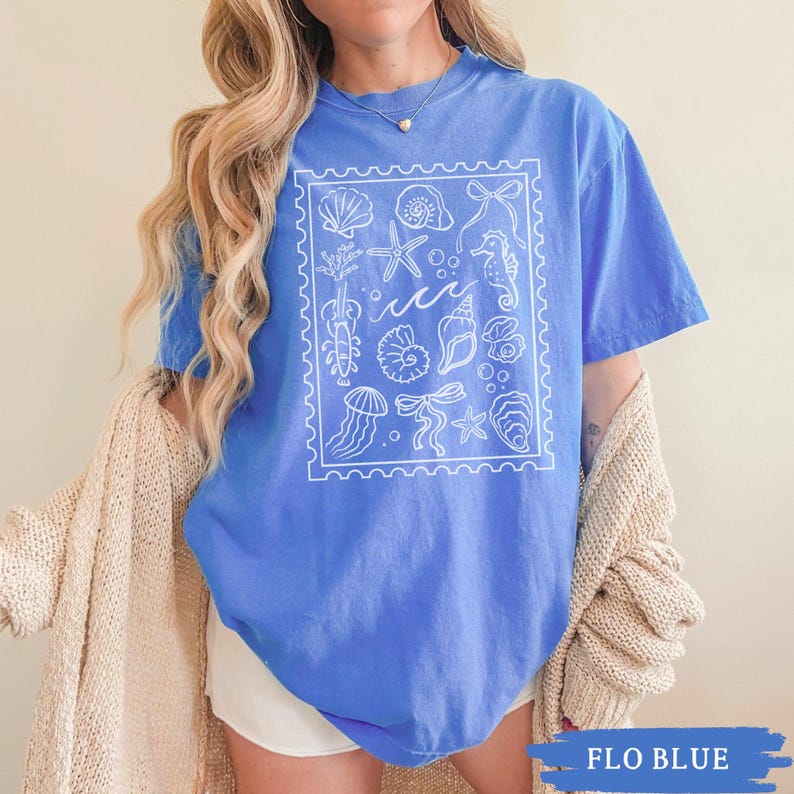 May include: A blue t-shirt with a white graphic of various sea creatures and shells inside a rectangular frame. The shirt has short sleeves and a crew neck. The text "FLO BLUE" is at the bottom of the image.