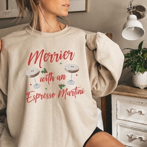 May include: A beige sweatshirt with the text "Merrier with an Espresso Martini" in red lettering. Two martini glasses with coffee are depicted on the sweatshirt, along with holly and red berries.