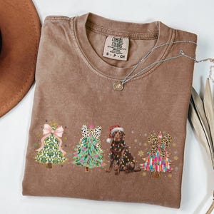 May include: A brown t-shirt with a Christmas-themed design featuring four decorated Christmas trees and a chocolate Labrador Retriever wearing a Santa hat. The trees are decorated with colourful ornaments, bows, and lights. The text "COMFORT COLORS" is visible on a tag attached to the shirt.