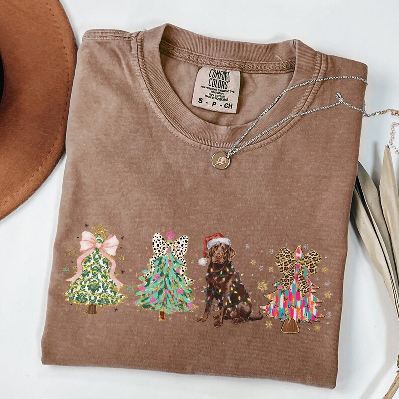 May include: A brown t-shirt with a Christmas-themed design featuring four decorated Christmas trees and a chocolate Labrador Retriever wearing a Santa hat. The trees are decorated with colourful ornaments, bows, and lights. The text "COMFORT COLORS" is visible on a tag attached to the shirt.