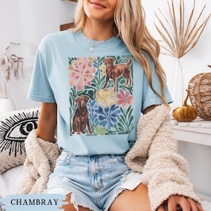 May include: Pale blue t-shirt with a watercolour illustration of two brown dogs and floral elements. The shirt has a loose fit and short sleeves. The word "CHAMBRAY" is printed at the bottom of the image.