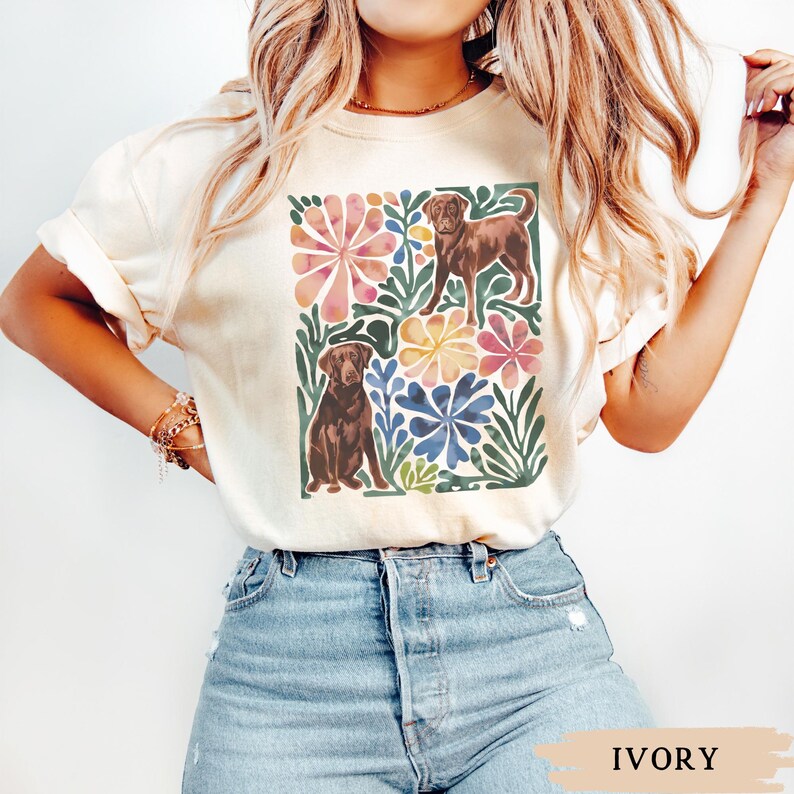 May include: Ivory t-shirt with a colourful floral design and two brown Labrador Retrievers. The design includes pink, yellow, and blue flowers with green foliage. The shirt is made of a soft material.