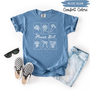 May include: A blue jean Comfort Colors t-shirt with a white floral design and the words "Flower Girl". The shirt is styled with sunglasses, checkered slip-on shoes, and denim shorts.