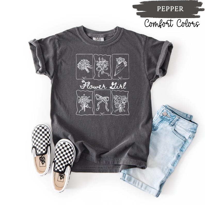 May include: A dark gray t-shirt with a white floral design and the text "Flower Girl." The shirt is paired with light blue denim shorts and black and white checkered slip-on shoes. The words "Pepper" and "Comfort Colors" are also visible.