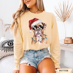 May include: A light yellow long-sleeve shirt featuring a graphic of a Boxer dog wearing sunglasses, a Santa hat, and a string of Christmas lights. The dog is blowing a bubblegum bubble. The word "BUTTER" is printed at the bottom.