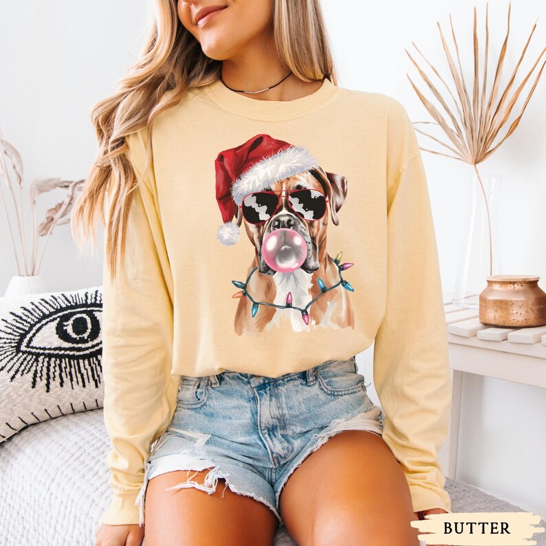 May include: A light yellow long-sleeve shirt featuring a graphic of a Boxer dog wearing sunglasses, a Santa hat, and a string of Christmas lights. The dog is blowing a bubblegum bubble. The word "BUTTER" is printed at the bottom.