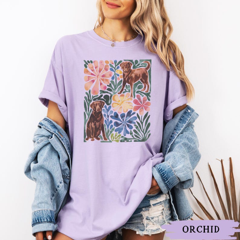May include: A lilac t-shirt with a watercolour design of two brown dogs surrounded by colourful flowers and foliage. The shirt has a relaxed fit and short sleeves. The word "ORCHID" is printed at the bottom of the image.