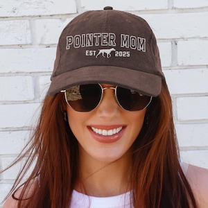 May include: A brown corduroy baseball cap with the text "POINTER MOM EST 2025" embroidered on the front. A white silhouette of a dog is also embroidered on the cap.