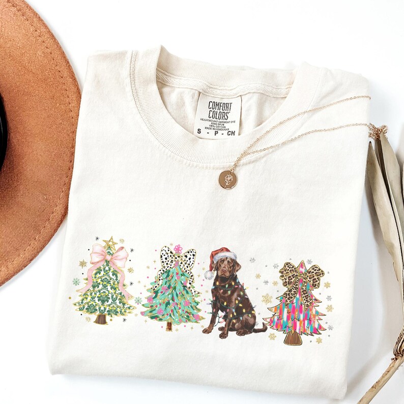 May include: A cream coloured t-shirt with a Christmas themed design featuring three decorated Christmas trees and a chocolate Labrador wearing a Santa hat. The text "COMFORT COLORS" is on the tag.
