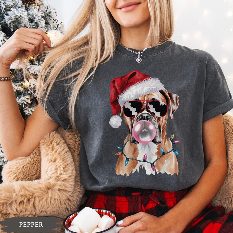 May include: Dark gray t-shirt featuring a Boxer dog wearing sunglasses and a Santa hat, blowing a bubblegum bubble. The dog is adorned with a string of Christmas lights. The word "PEPPER" is at the bottom.
