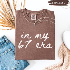 May include: A brown t-shirt with the text "in my 67 era" in white script. The shirt is folded and has a silver necklace with a sun pendant. A pair of blue jeans and a white sneaker are also in the image.