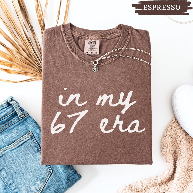 May include: A brown t-shirt with the text "in my 67 era" in white script. The shirt is folded and has a silver necklace with a sun pendant. A pair of blue jeans and a white sneaker are also in the image.