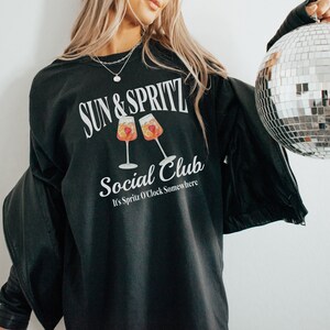 May include: Black sweatshirt with white text that reads "Sun & Spritz Social Club It's Spritz O'Clock Somewhere". The text is surrounded by a graphic of two glasses of orange juice with fruit.