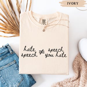 May include: Ivory-colored t-shirt with the text "hate speech &ne; speech you hate" in black script. The shirt is folded and accessorized with a delicate necklace. A pair of blue jeans and a white shoe are visible in the background.