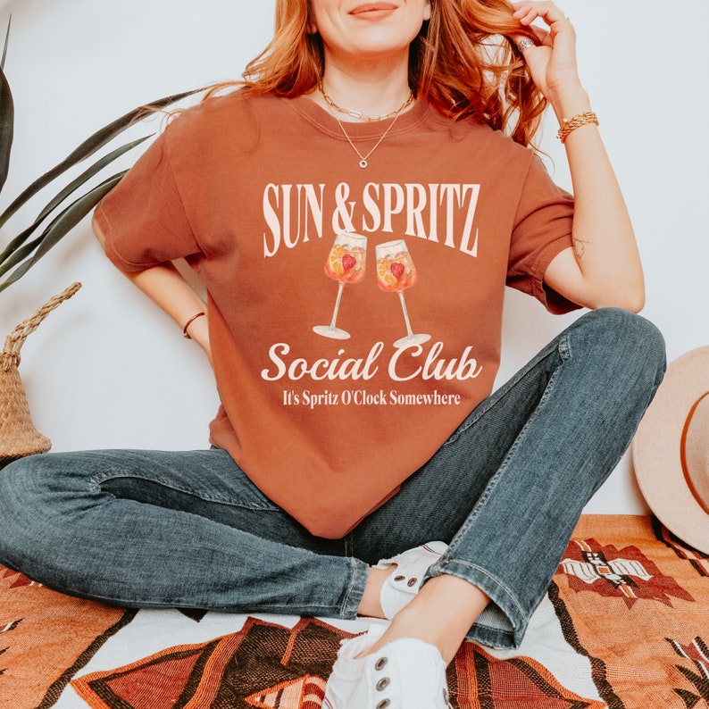 May include: A rust-colored t-shirt with the text "Sun & Spritz Social Club" and the phrase "It's Spritz O'Clock Somewhere" in white. The shirt features a graphic of two glasses of spritz with fruit.