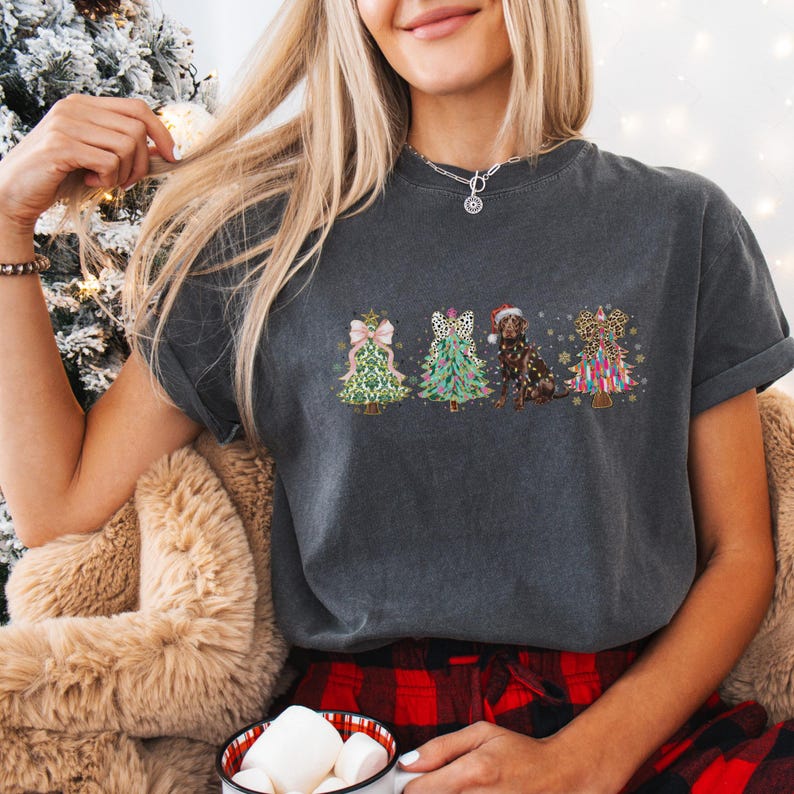May include: A dark grey t-shirt with a Christmas tree design featuring a dog and bows. The trees are decorated with different patterns, including leopard print, polka dots and plaid.
